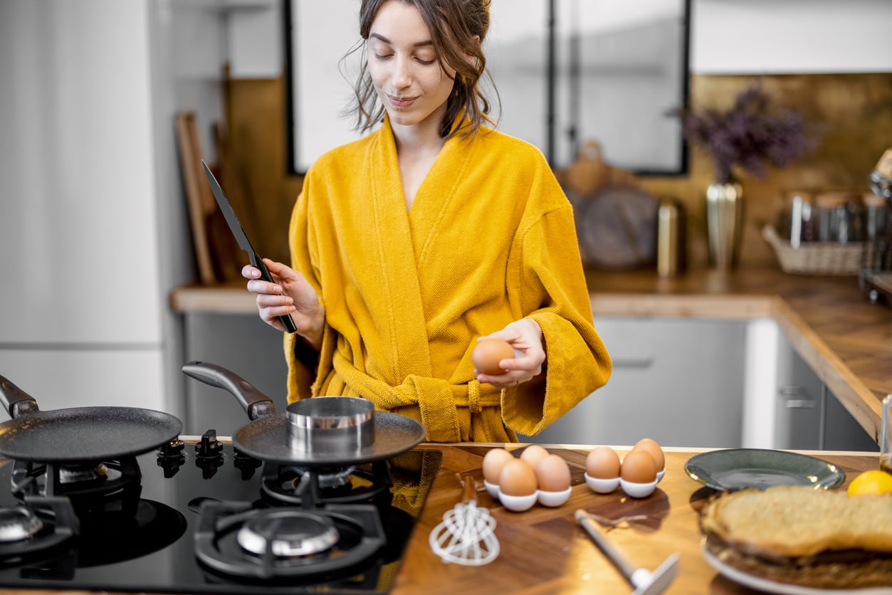 Woman in a yellow robe stands in a kitchen holding an egg and a knife, preparing to cook at a stove. Eggs, a whisk, and a stack of crepes—resembling delicious foods to order at your favorite café—are on the wooden counter beside her.
