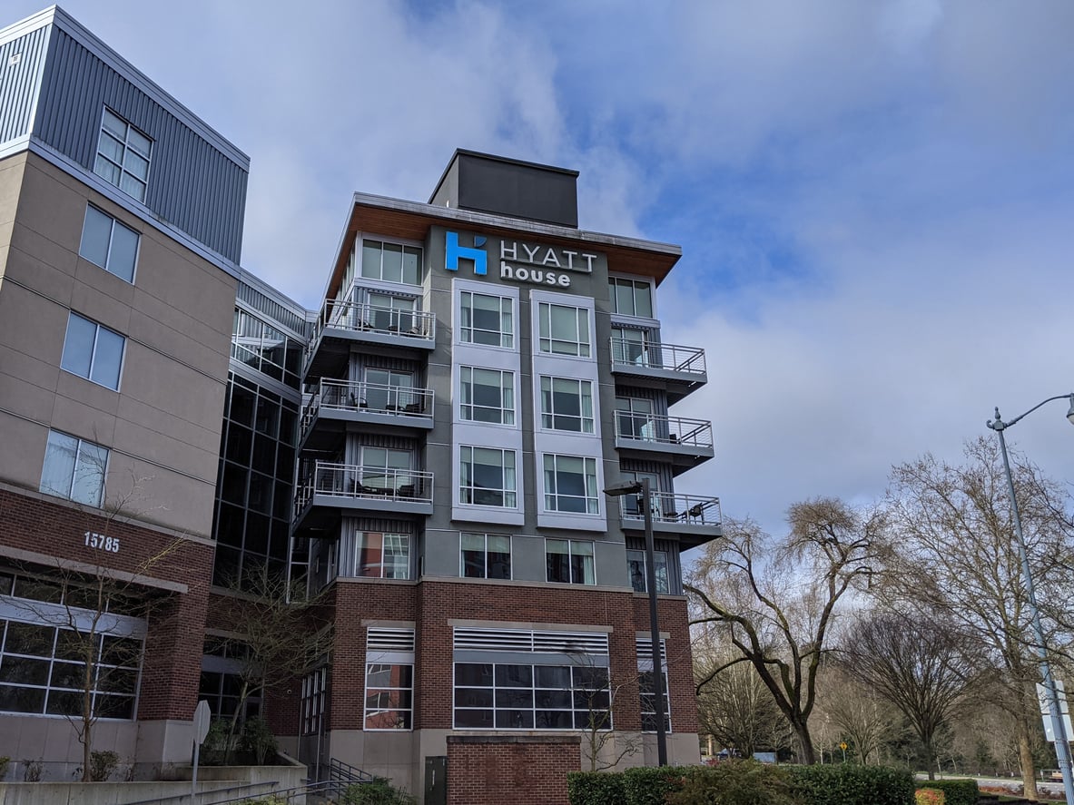 A modern multi-story Hyatt House hotel building with balconies, large windows, and the blue hotel sign—representing one of the premier hotel chains. Trees and a cloudy blue sky are in the background. The address number 15785 is visible on the building.