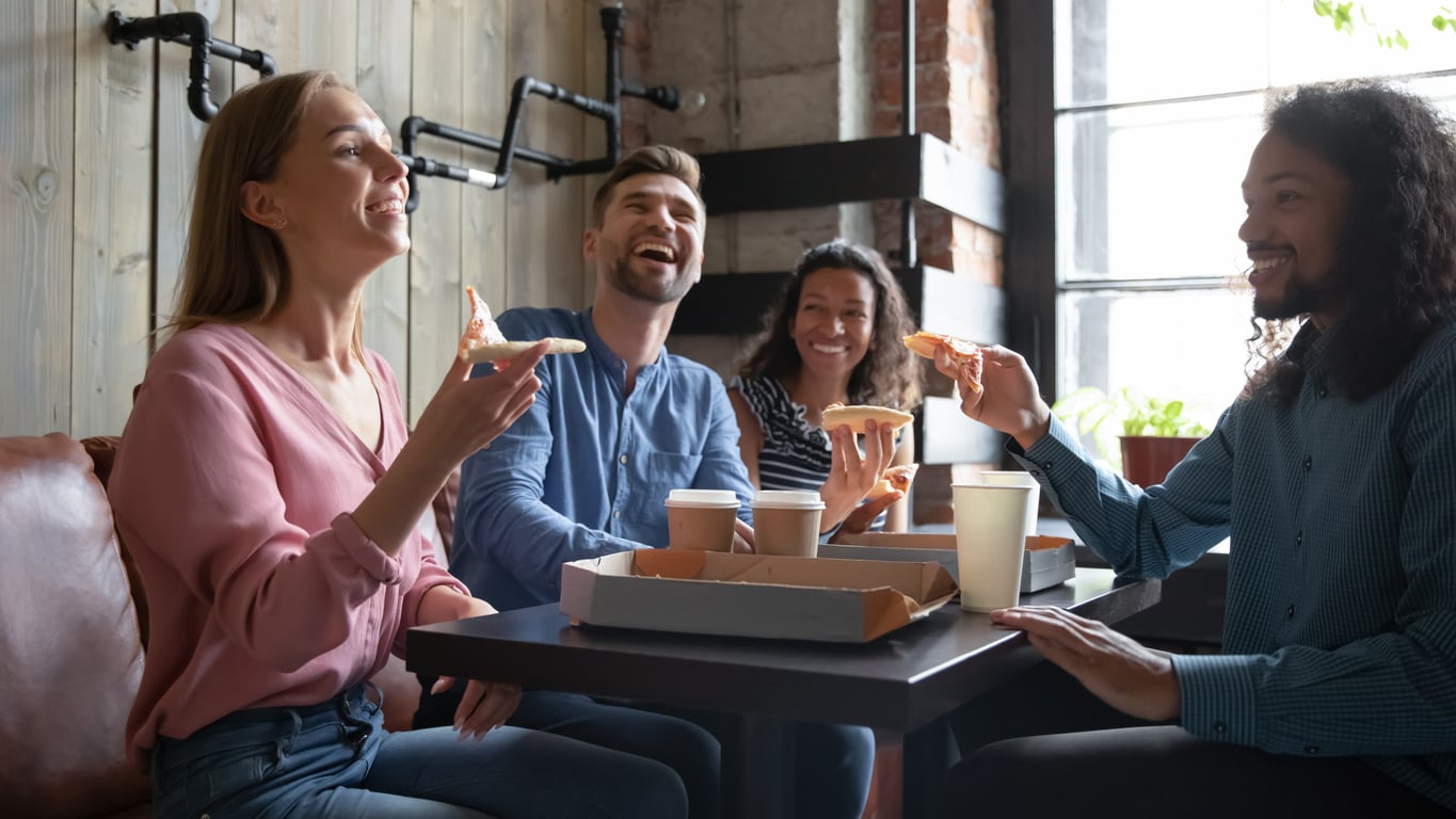 Four people sit around a table, smiling and laughing while eating pizza and drinking coffee. Sunlight streams in through a window, creating a warm, casual atmosphere.