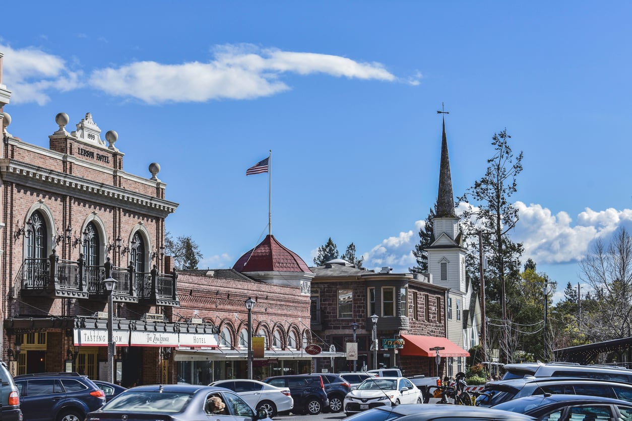 Historic downtown street with brick buildings, storefronts, a church steeple, American flag, trees, and parked cars under a blue sky with clouds.