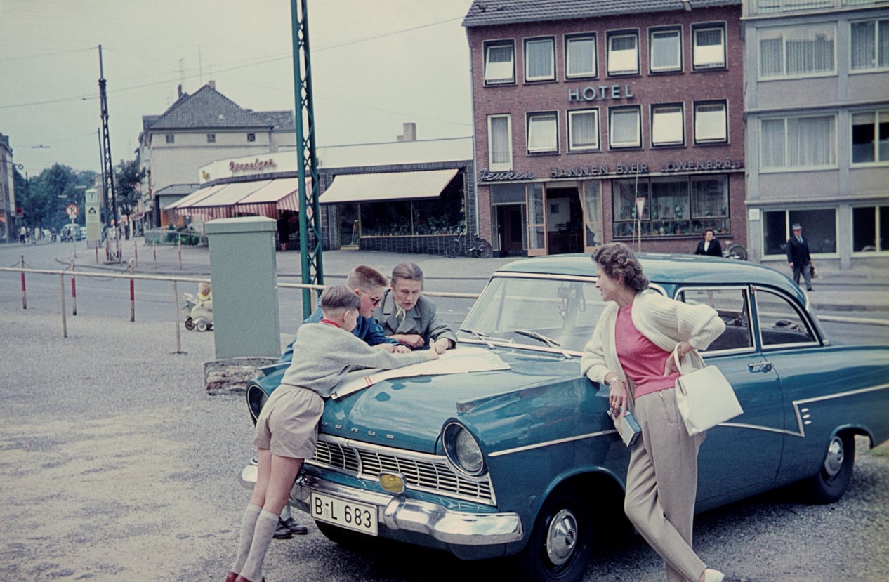Two boys and a man look at a map on the hood of a blue vintage car while a woman stands nearby. Reminiscent of travel in the 60s and 70s, they are parked near a hotel and shops on a city street.