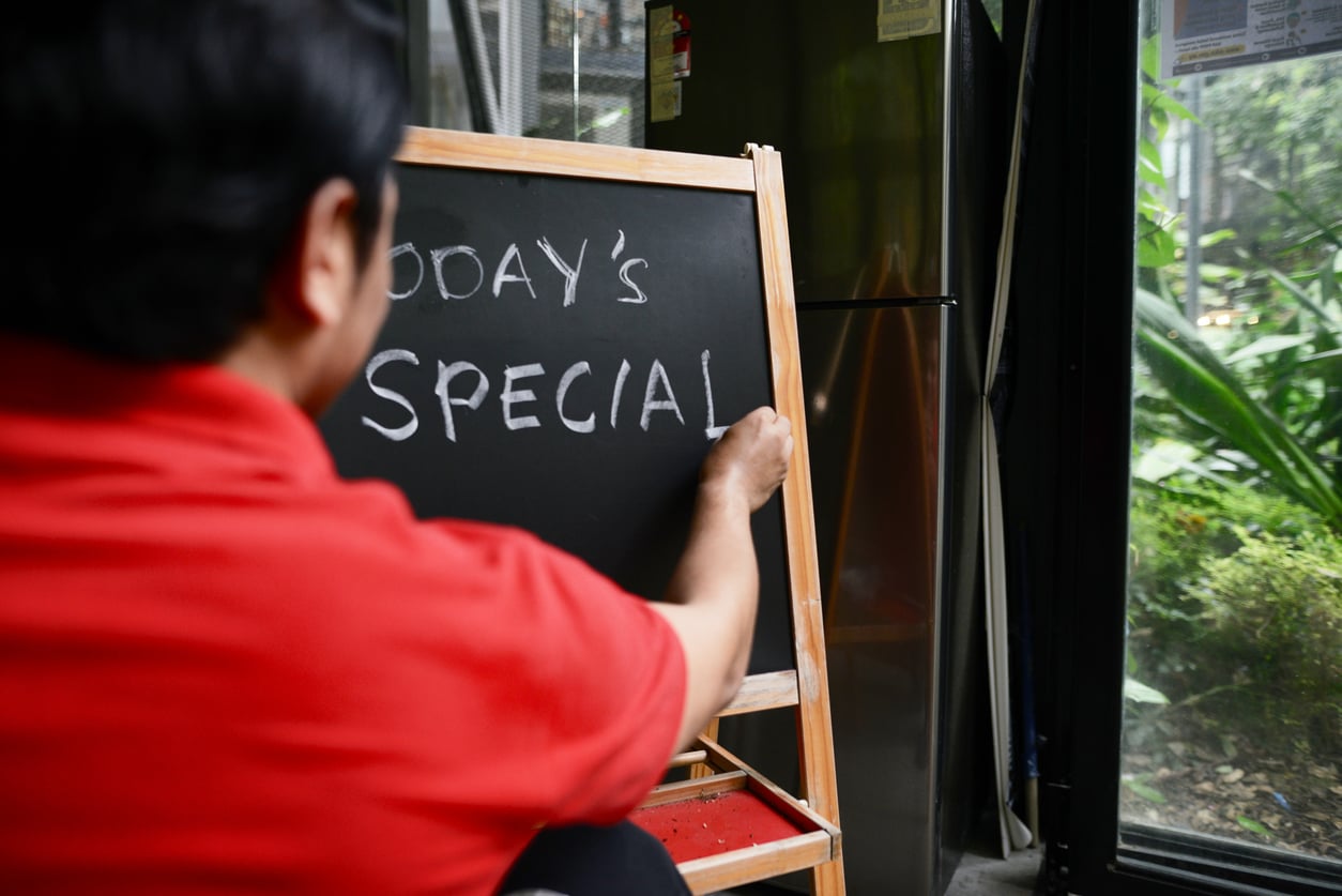 A person in a red shirt writes “TODAY’S SPECIAL” on a small black chalkboard inside a cafe which is one of the restaurant trends, near a window with green plants visible outside.