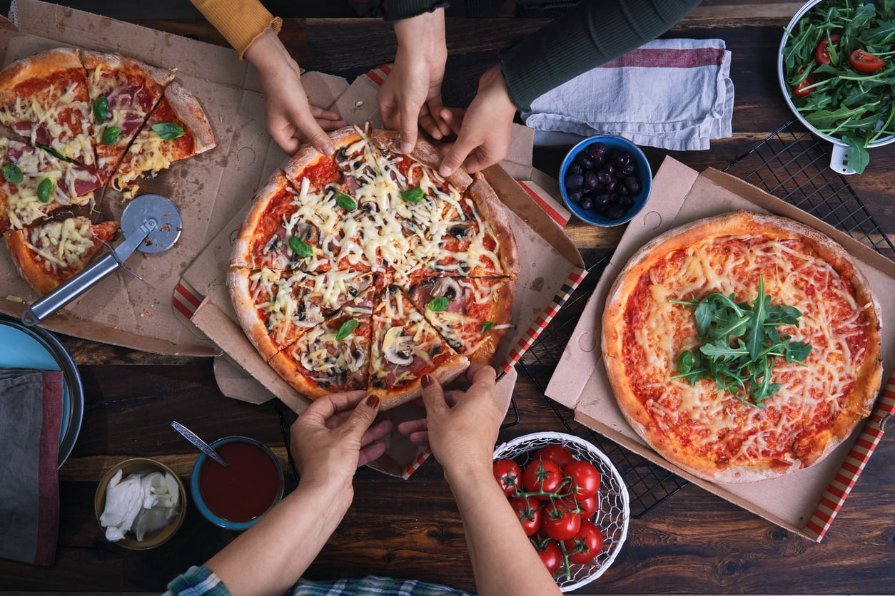 Three people share slices from two pizzas on a wooden table, surrounded by fresh ingredients like tomatoes, arugula, and olives. A third pizza, topped with arugula, remains untouched in the foreground.