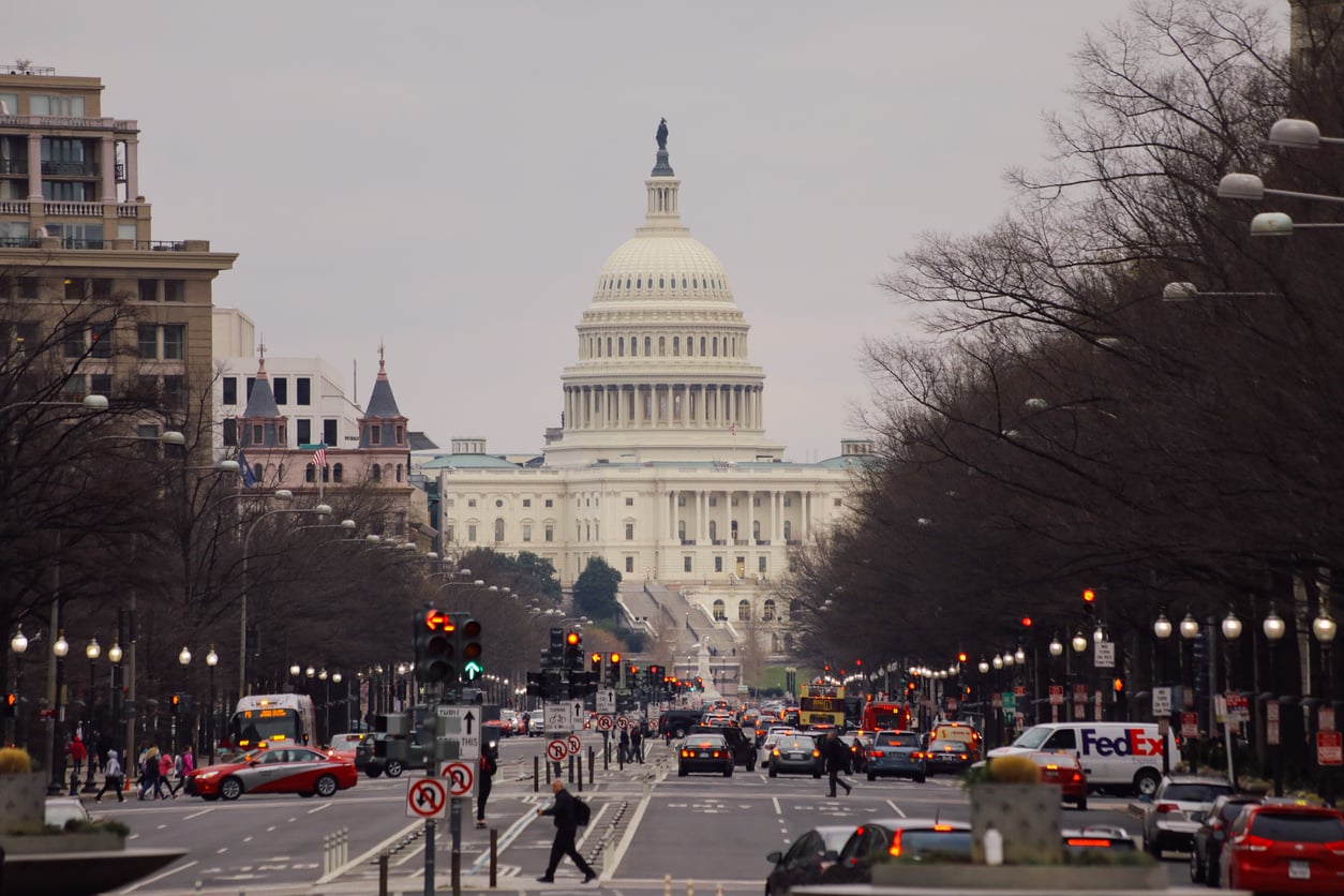 The U.S. Capitol building stands in the background, framed by city buildings, leafless trees, and a busy street filled with cars, buses, and pedestrians on a cloudy day.