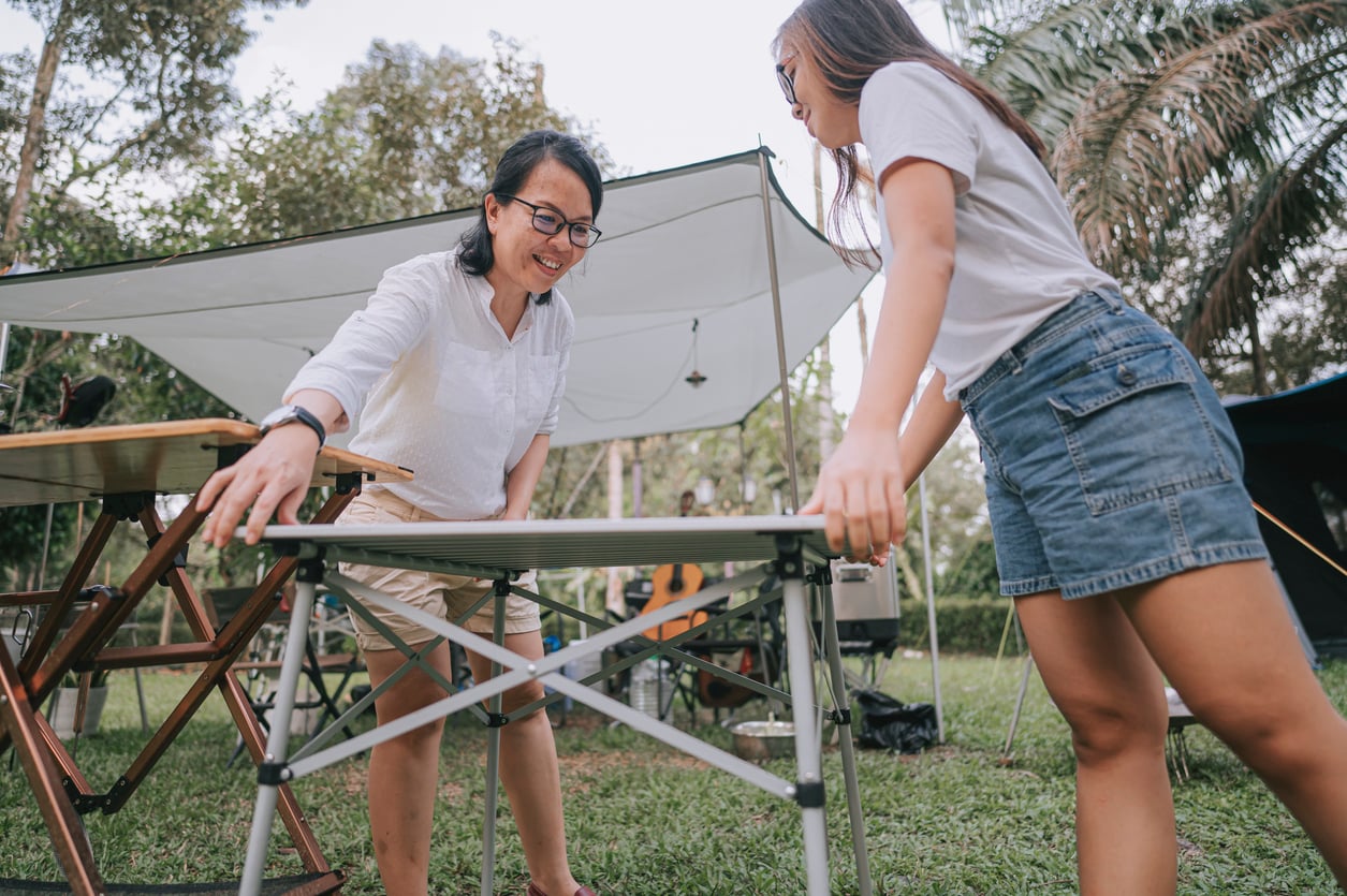 Two people set up a folding table at a campsite, smiling and working together. There are trees, grass, and other camping equipment visible in the background.