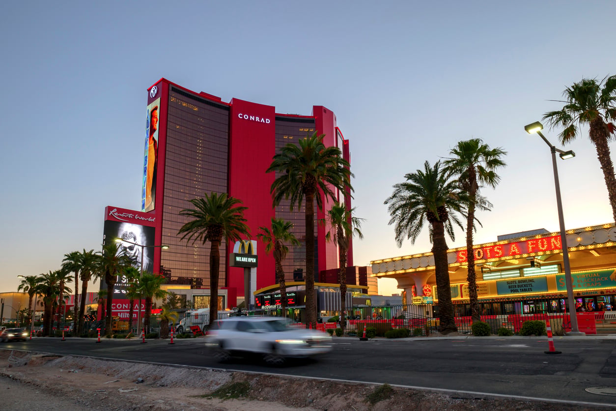 A white truck drives past palm trees, Resorts World hotel, and a lit “SLOTS A FUN” casino sign in Las Vegas at dusk.