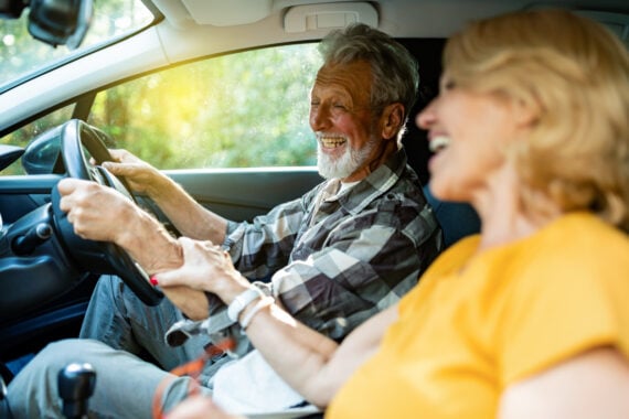 An older man and woman laugh together inside a car, with the man in the driver&rsquo;s seat holding the steering wheel and the woman in the passenger seat, both appearing joyful and relaxed.
