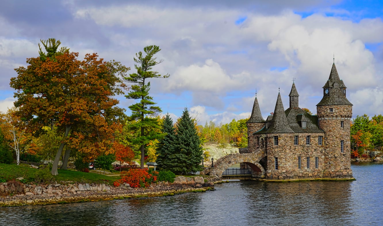 A majestic stone castle with pointed towers sits by the water, surrounded by autumn trees with colorful foliage under a partly cloudy sky—a timeless addition to the legacy of grand castles in picturesque landscapes.