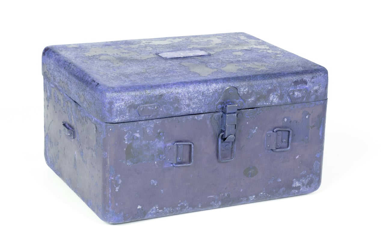 A weathered, rectangular blue metal box with a latch and two side handles, showing signs of rust and wear, set against a white background.