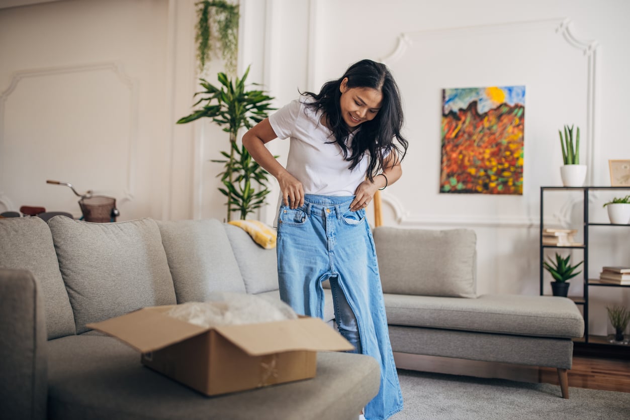 A woman smiles while holding up a pair of jeans in front of her, standing in a living room with an open cardboard box on the couch.