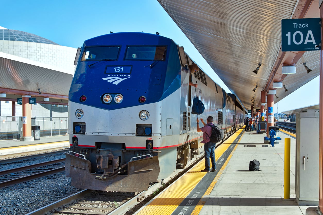 A silver and blue Amtrak train is stopped at a station platform. A person with a backpack stands next to the train, and a sign reads “Track 10A.” The sun casts sharp shadows on the ground.