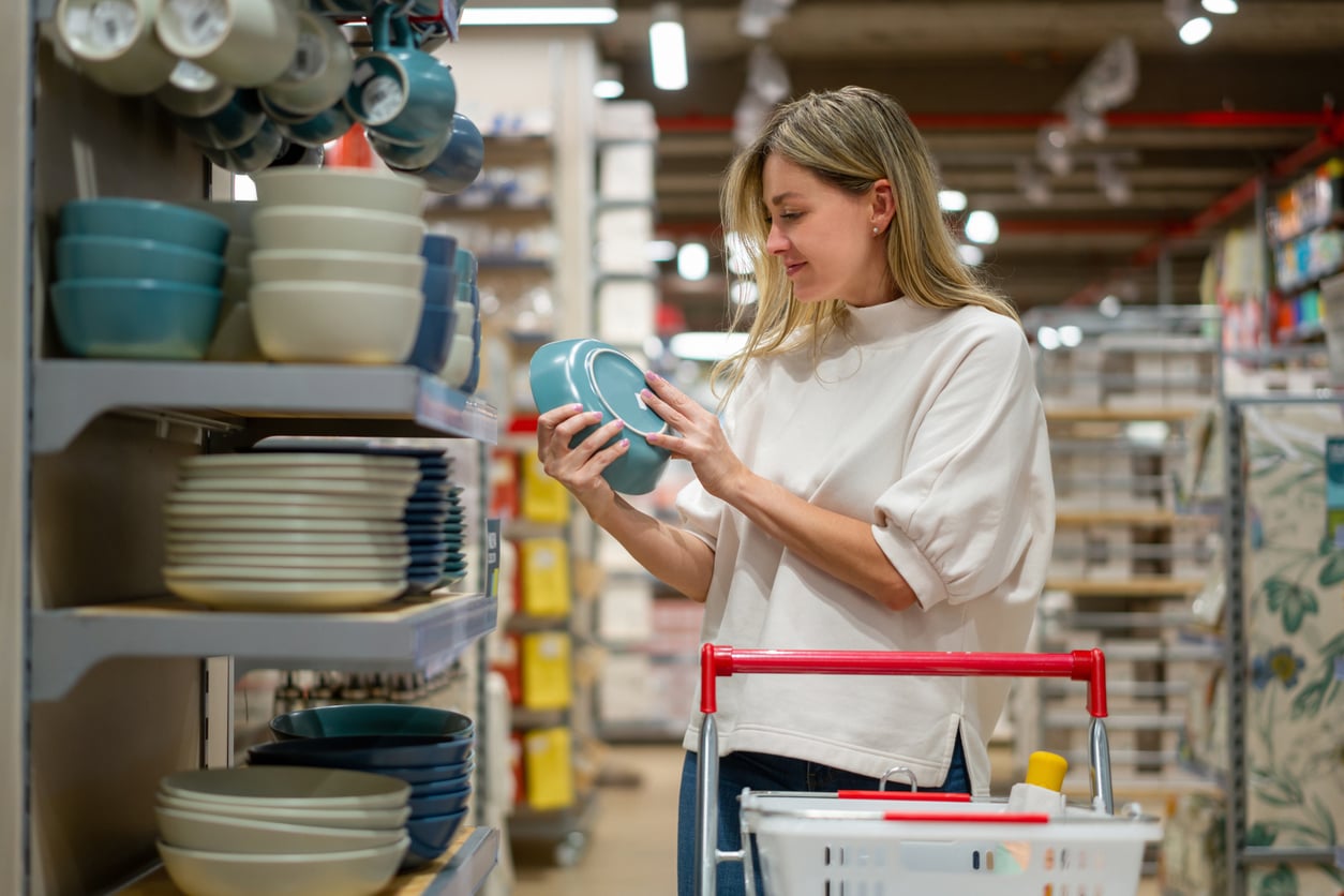 A woman stands in a store aisle, holding and examining a blue bowl. She is next to shelves filled with stacked dishes, with a shopping cart containing items beside her.