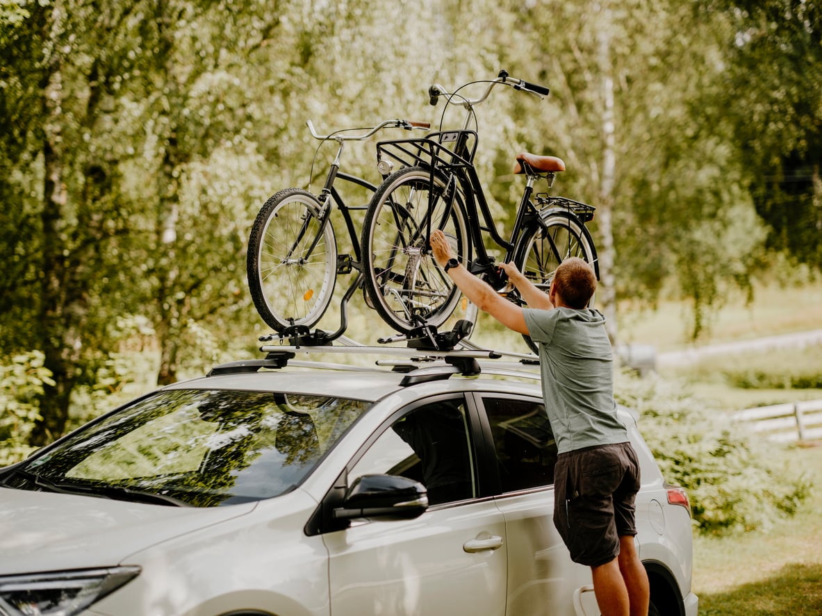 A person secures two bicycles onto the roof rack of a white car parked outdoors, surrounded by green trees and nature.