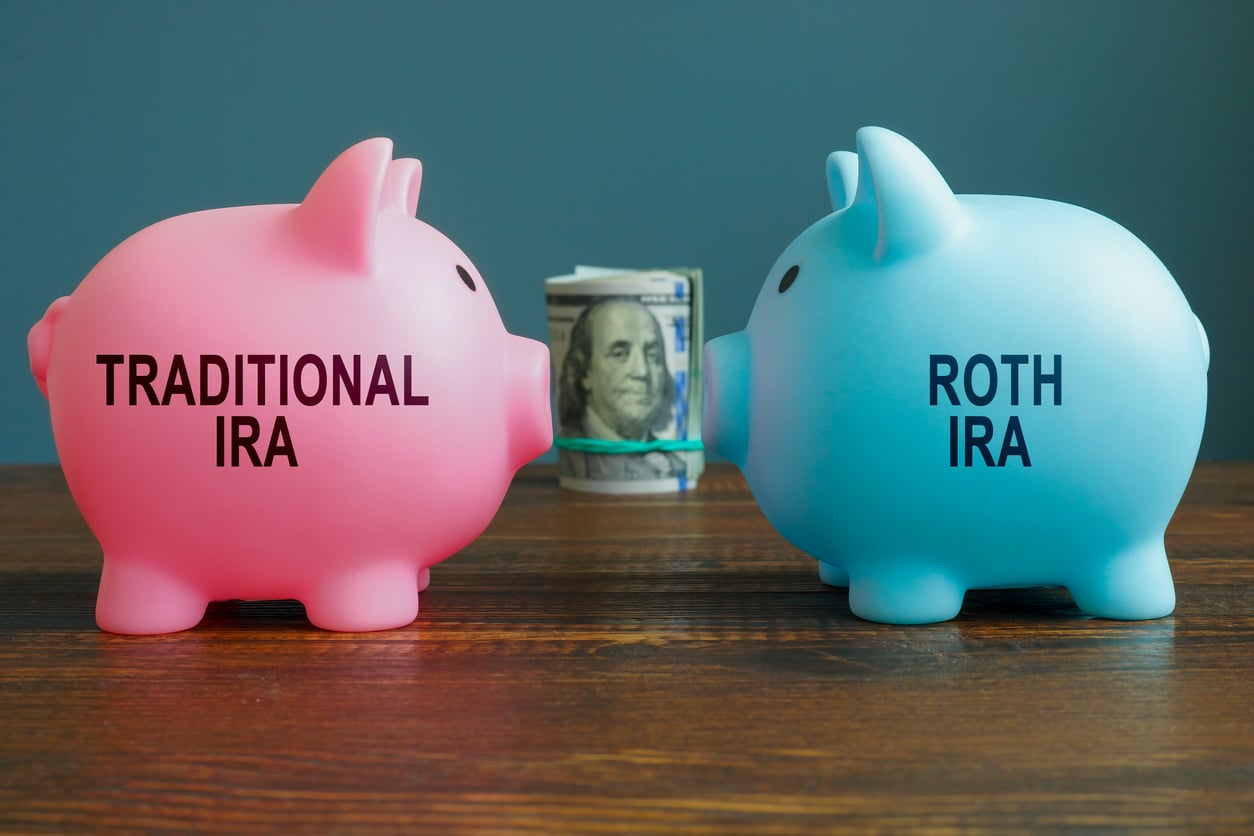 Two piggy banks, one pink labeled “Traditional IRA” and one blue labeled “Roth IRA,” face each other with a rolled-up $100 bill between them on a wooden table, against a plain blue background.