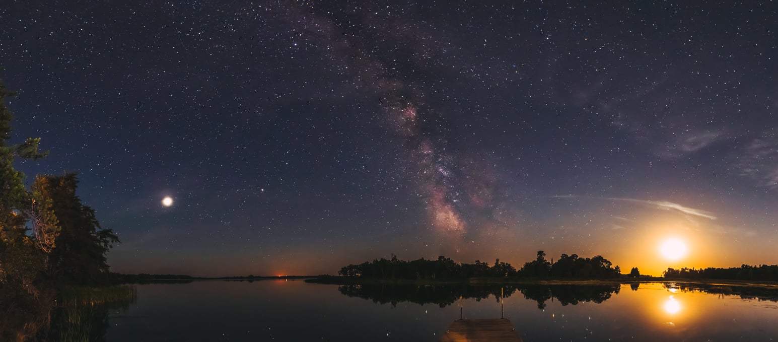 A star-filled night sky with the Milky Way visible, reflected in a calm lake. The moon and another bright celestial object shine over a silhouetted treeline and dock.