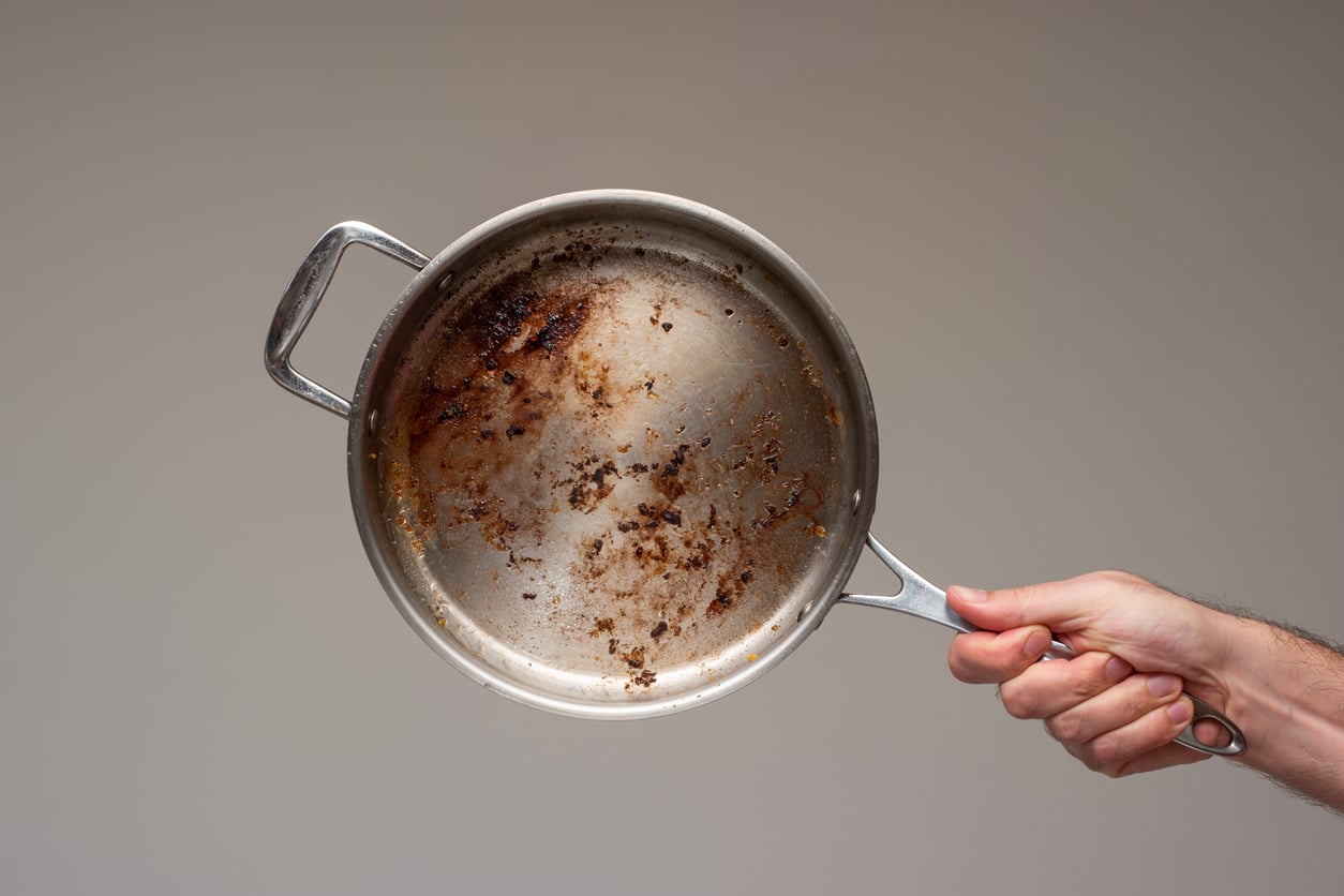 A hand holds a stainless steel pan with burnt food residue stuck to the bottom against a plain light background.