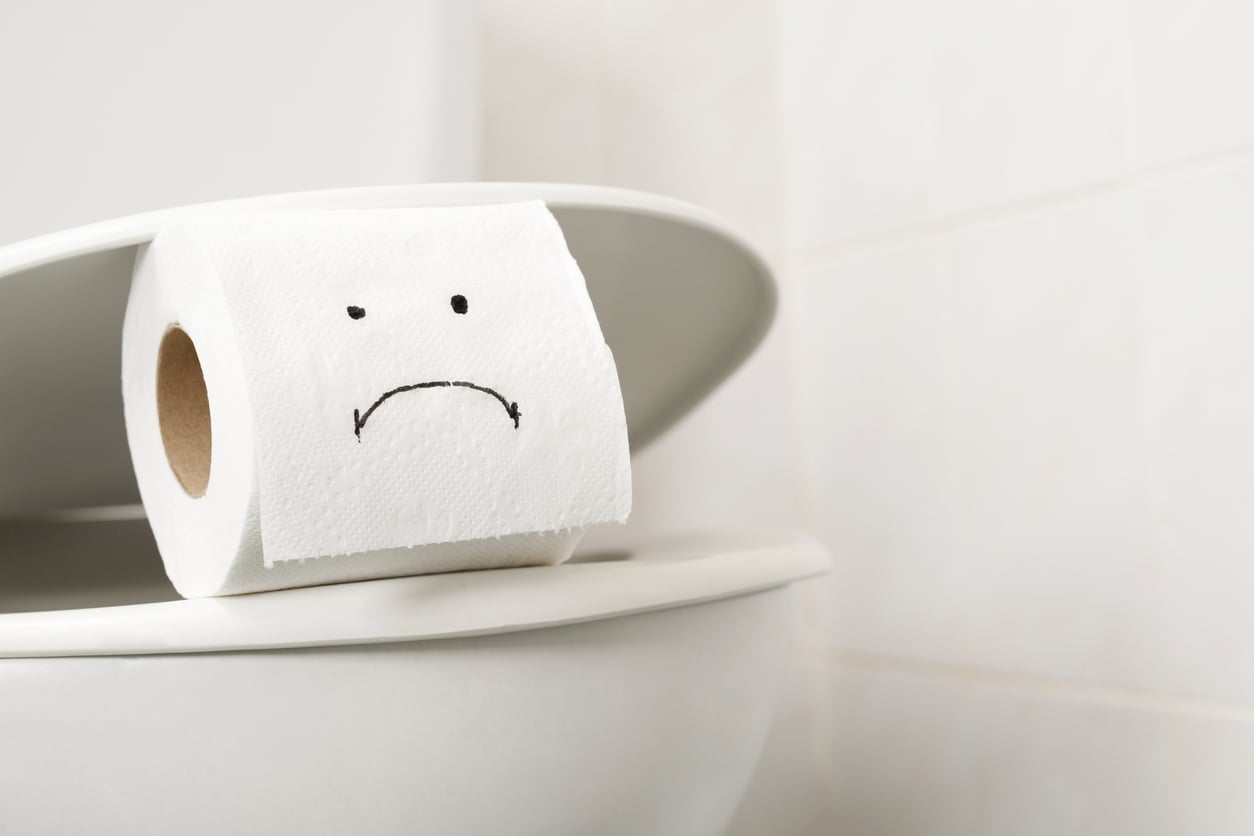 A toilet paper roll with a sad face drawn on it sits on top of a toilet lid in a white bathroom.