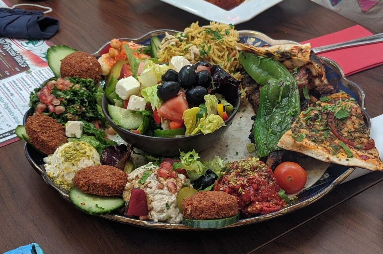A platter of Mediterranean appetizers including falafel, tabbouleh, hummus, salad with feta and olives, baba ghanoush, grape leaves, pita bread, and slices of flatbread, all arranged on a decorative plate.