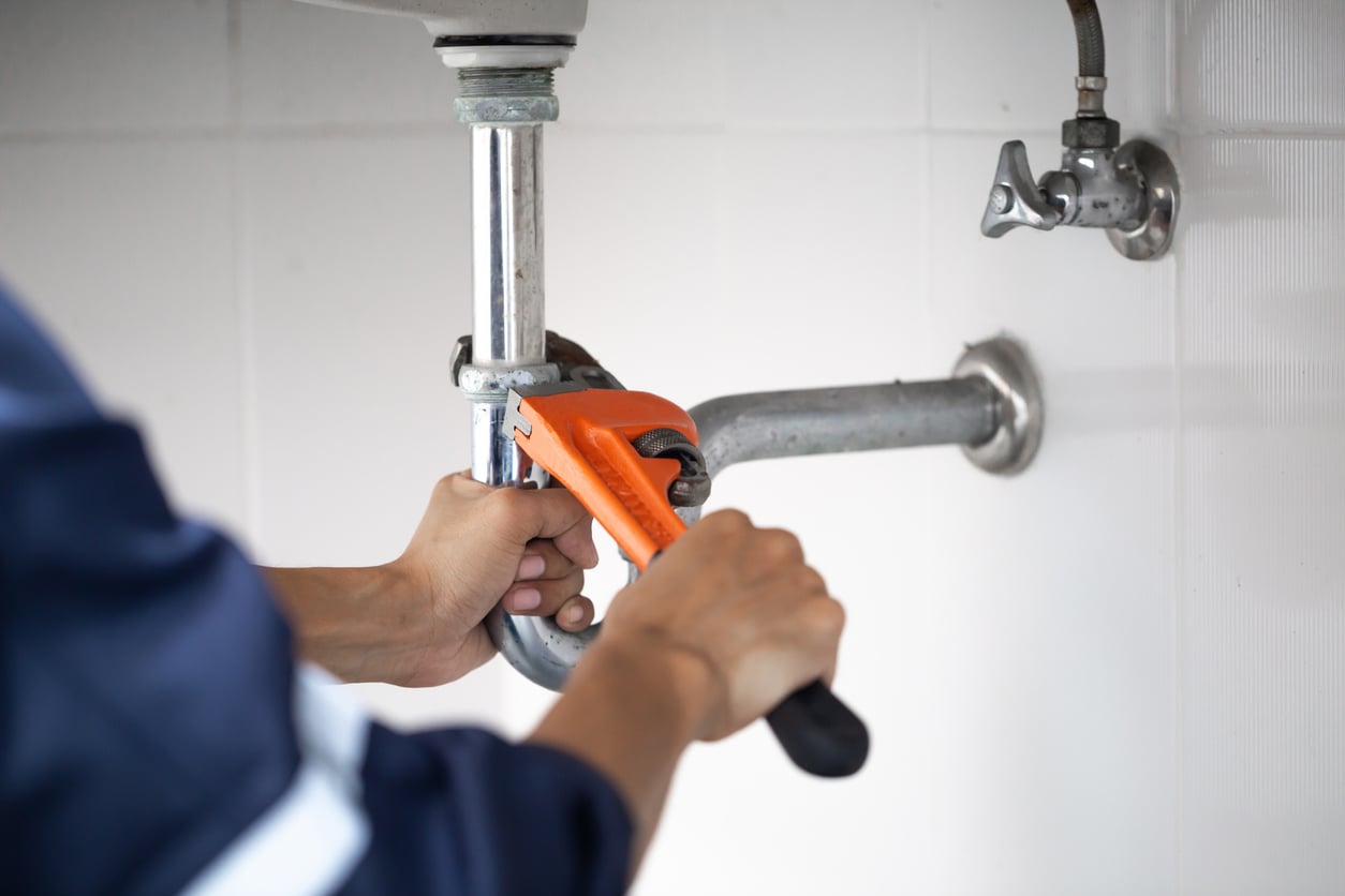 Person using an orange pipe wrench to tighten or loosen a pipe under a sink against a white tiled wall.
