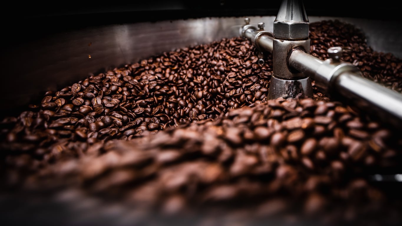 Close-up of roasted coffee beans being stirred inside a large metal roasting machine, with shiny metal arms moving through the beans.