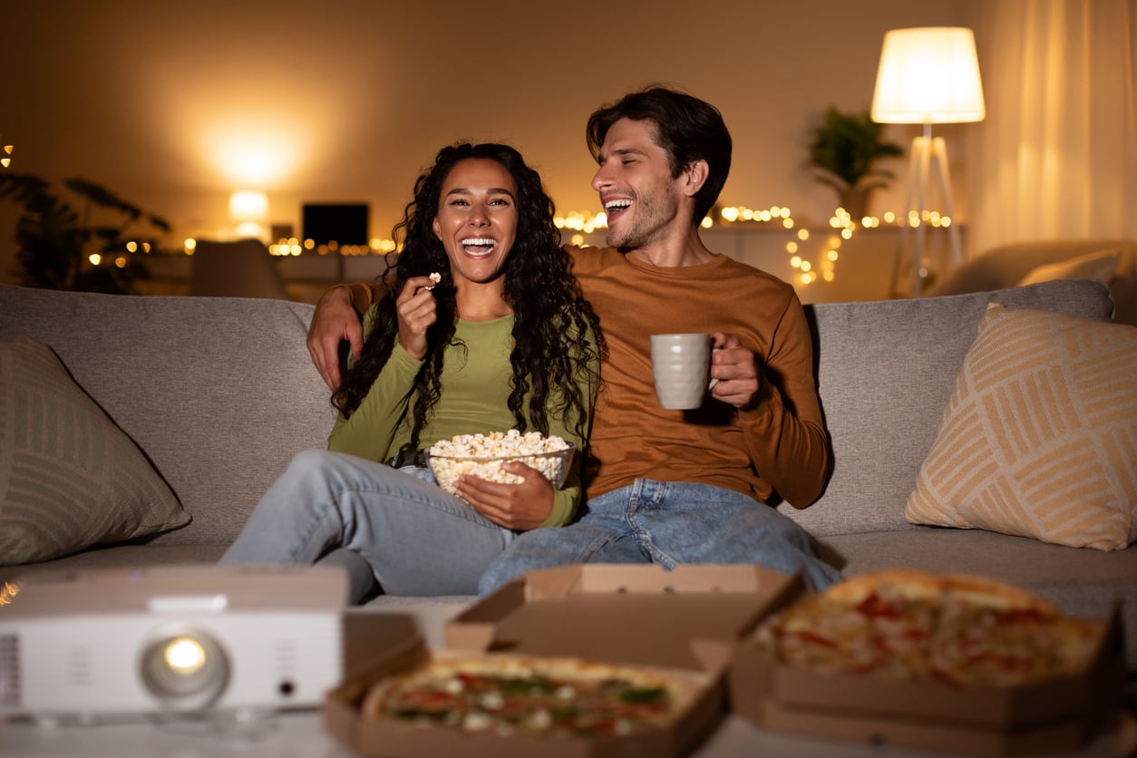 A couple sits on a couch, laughing and eating popcorn. Pizza boxes are on the table in front, and a projector is nearby. Warm lighting and string lights decorate the cozy living room.