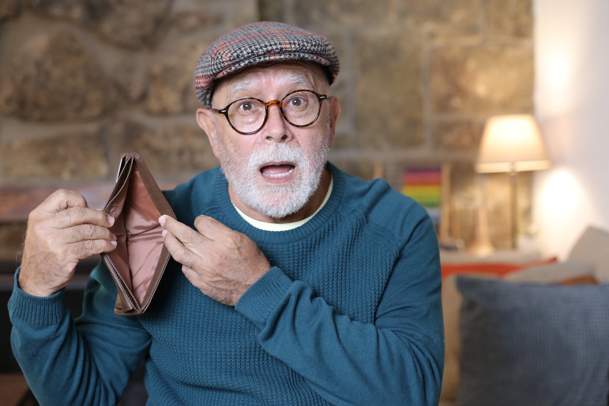 An older man wearing glasses, a cap, and a teal sweater looks surprised while holding open an empty wallet in a cozy living room setting.