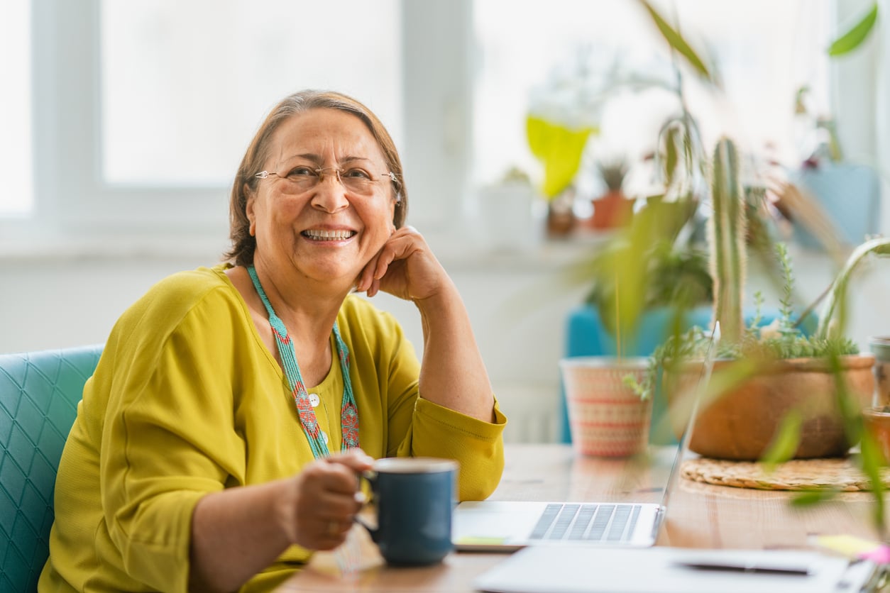 An older woman in a yellow top smiles while holding a mug at a desk with a laptop, surrounded by houseplants in a bright, cozy room—perhaps reflecting on her retirement money habits as she enjoys this peaceful moment.