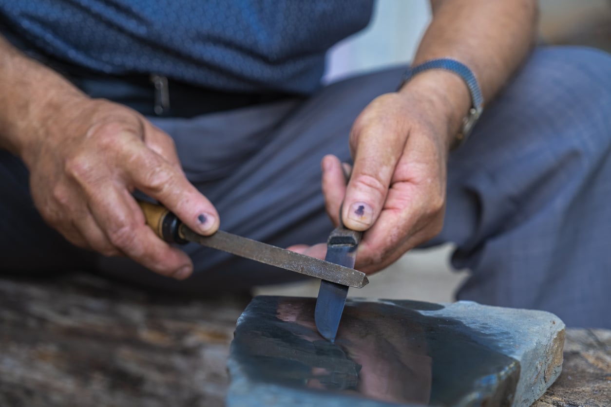 A person sharpens a knife on a sharpening stone, holding the blade in one hand and a tool in the other. The surface below is rough wood. The person wears a blue shirt and gray pants.