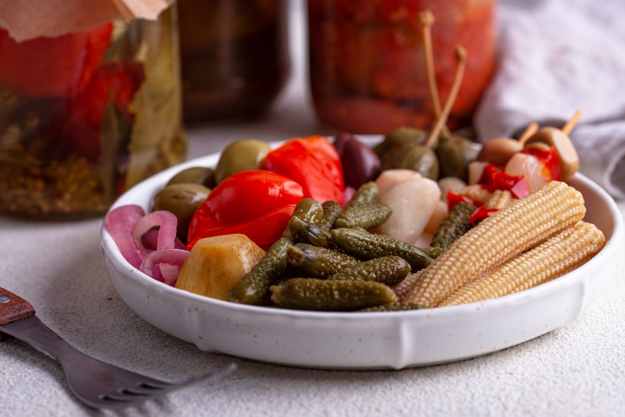 A white plate with assorted pickled vegetables including gherkins, baby corn, olives, onions, red peppers, and carrots. Jars of pickles and a fork are in the background.