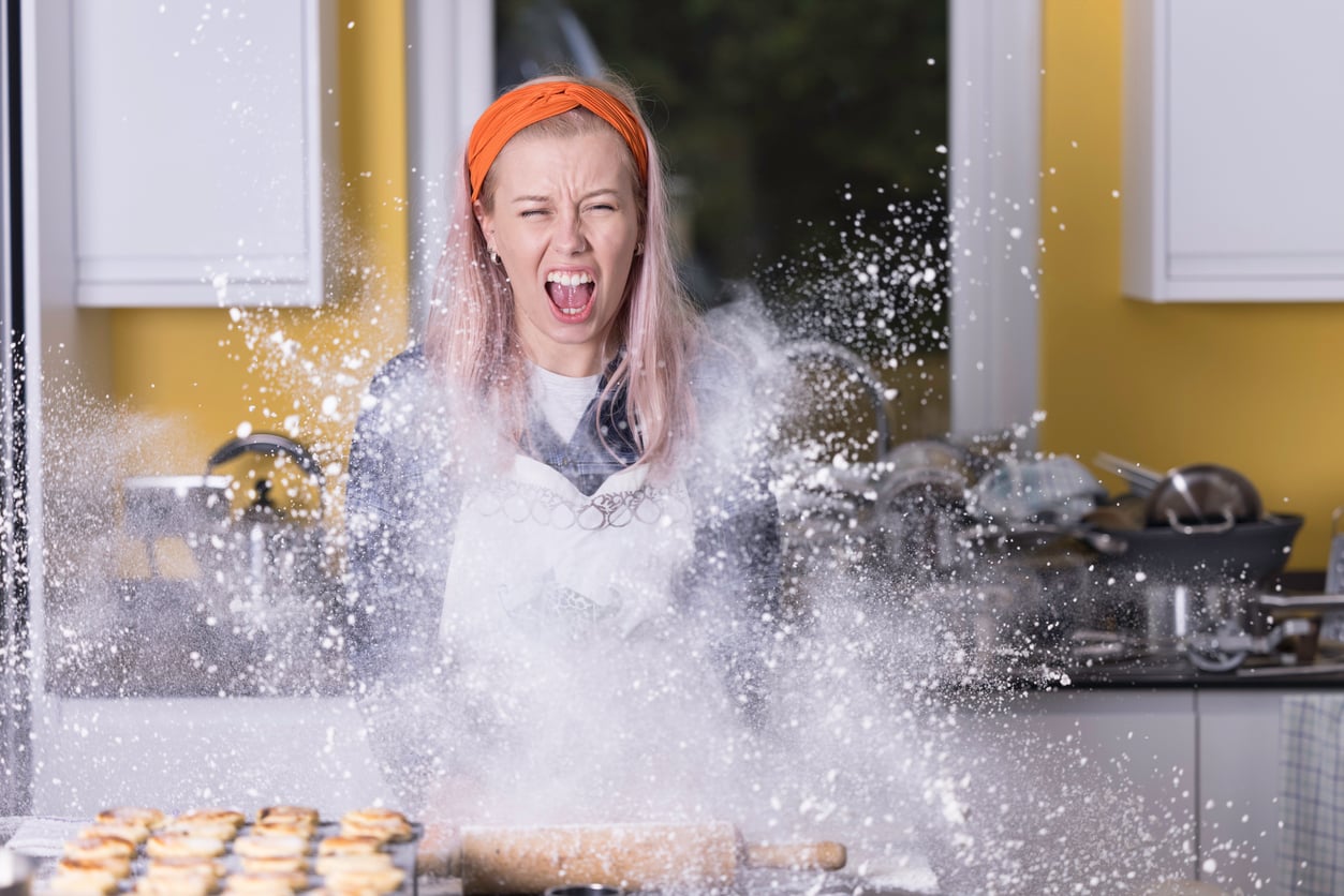 A woman in a kitchen wearing an orange headband and white apron yells as a cloud of flour explodes in front of her, with baked goods and a rolling pin on the counter.