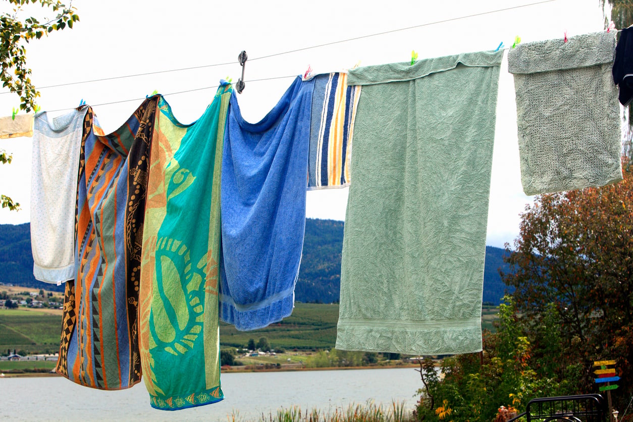 Colorful towels and a washcloth hang on a clothesline outdoors, with a lake, green hills, and trees visible in the background.