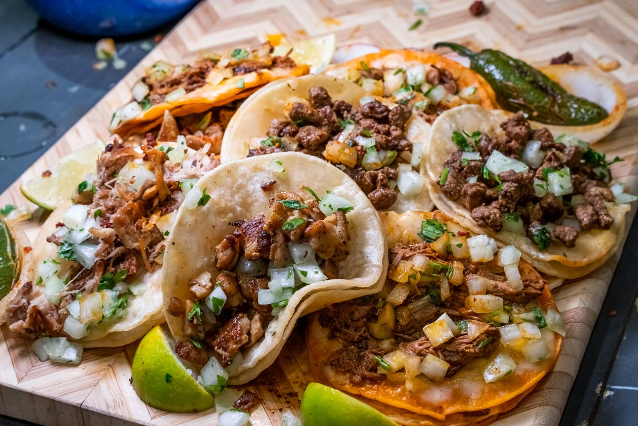 A wooden board filled with various tacos topped with diced onions, cilantro, and meat, alongside lime wedges and a roasted jalapeño.