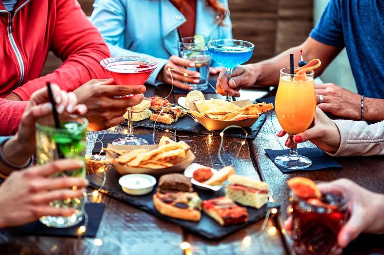 A group of people sit around a table, holding colorful cocktails and sharing appetizers, with string lights on the wooden surface, creating a festive and relaxed atmosphere.
