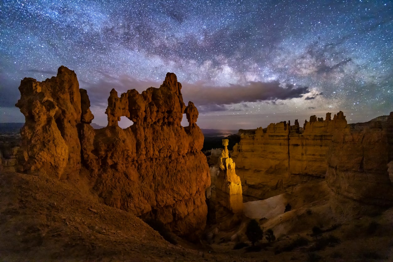 Rocky hoodoo formations are illuminated under a star-filled night sky and visible Milky Way, with dramatic clouds and rugged terrain in the foreground and background.