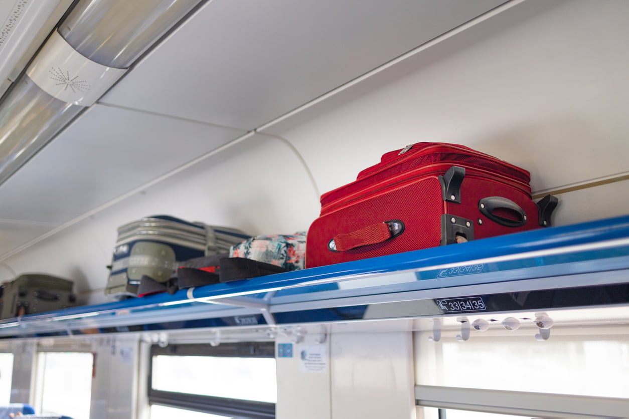 Several suitcases, including a prominent red one, are placed on a blue overhead luggage rack inside a train carriage.