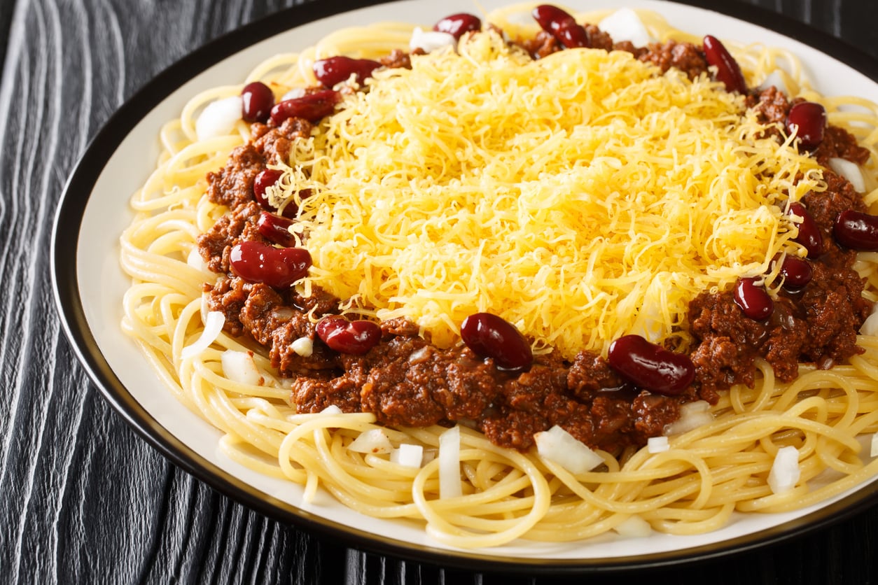A plate of spaghetti topped with chili, kidney beans, chopped onions, and a generous layer of shredded yellow cheese on a black and white plate, set on a dark wooden table.