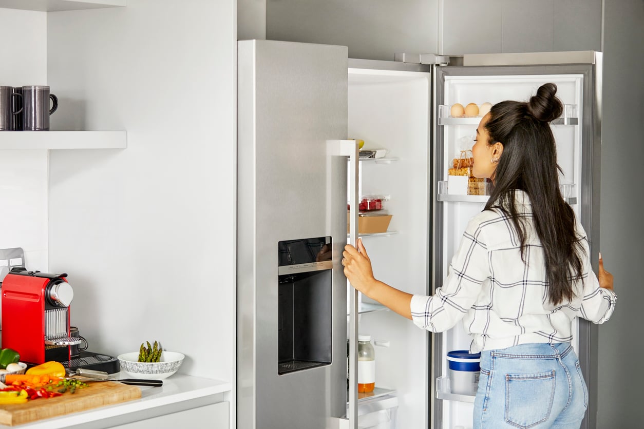 A woman in a plaid shirt opens a refrigerator door in a modern kitchen, looking inside—perhaps considering her grocery habits as she surveys the contents. Fresh vegetables and a red coffee machine are visible on the counter nearby.