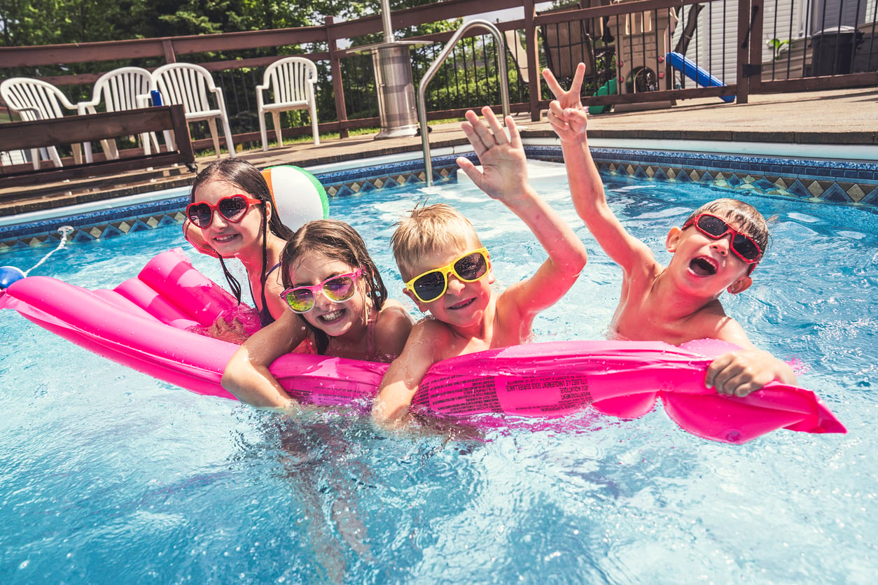 Four smiling children wearing sunglasses play on a pink pool float in a swimming pool, waving and making peace signs, capturing the carefree spirit of vacation, with pool chairs and a wooden deck in the background.