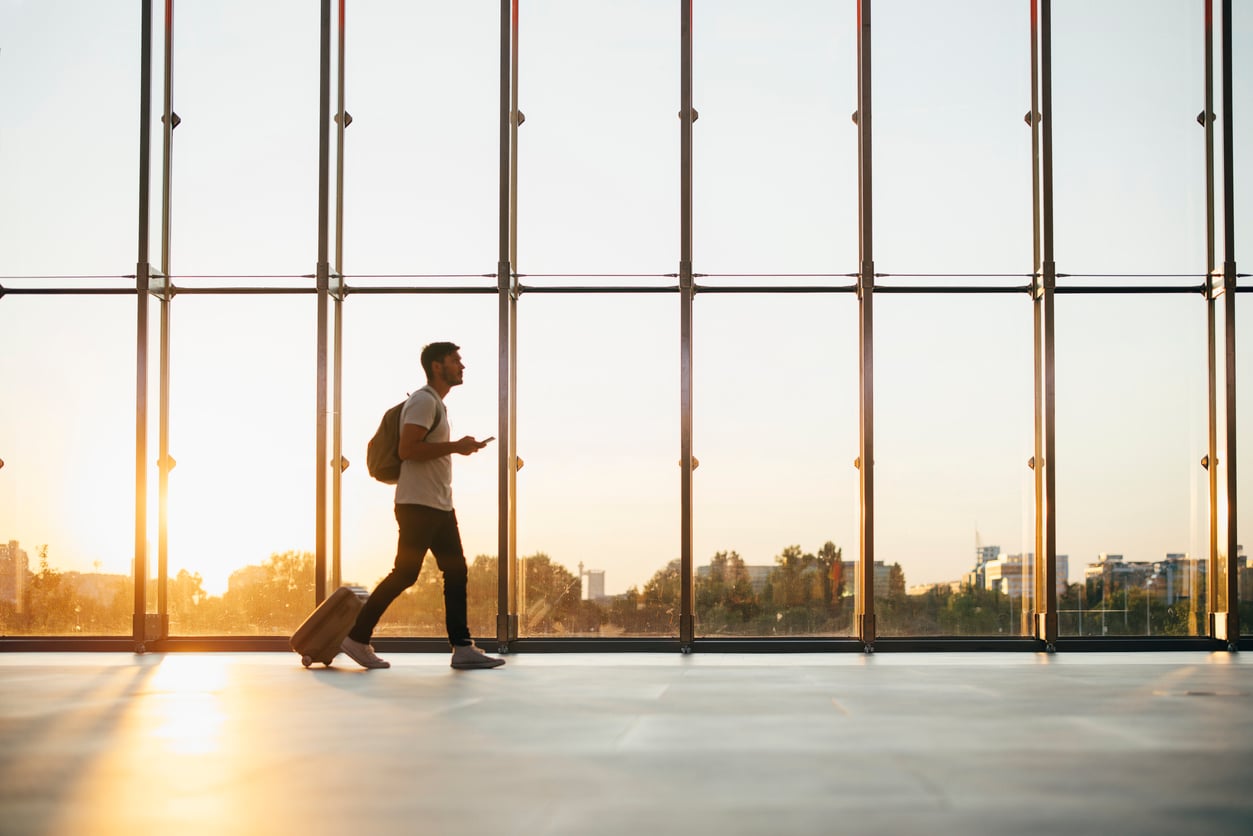 A person with a backpack and suitcase walks through an airport terminal at sunrise or sunset, silhouetted against large windows with a cityscape and trees visible outside—perhaps reflecting on past vacation mistakes as they embark on their next adventure.