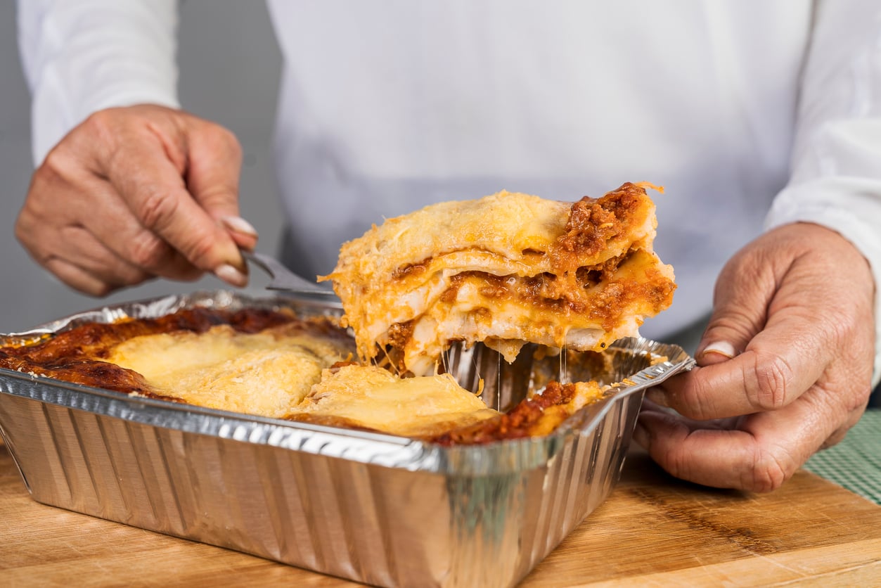 A person serves a cheesy slice of lasagna from a foil tray, showing layers of pasta, sauce, and melted cheese—one of the classic foods to order when craving comfort and flavor.