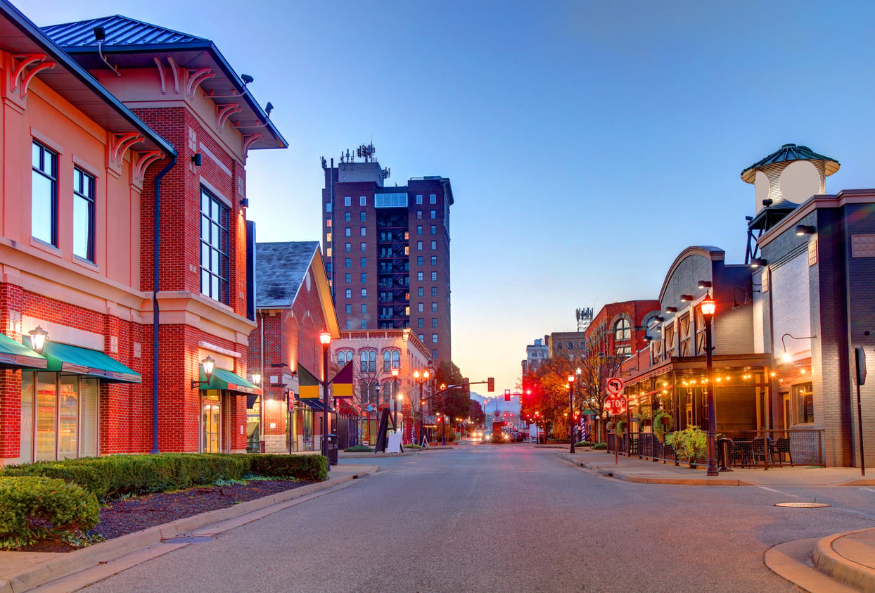 A quiet city street at sunset features brick buildings, storefronts with green awnings, streetlights, and a tall building in the background under a clear sky.