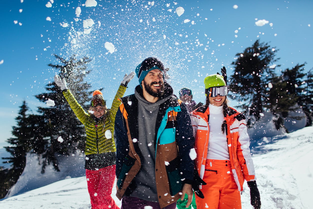 Three people in colorful winter gear walk through snow, smiling and laughing as snow is thrown into the air. Snow-covered trees and a bright blue sky are in the background of the winter vacation.