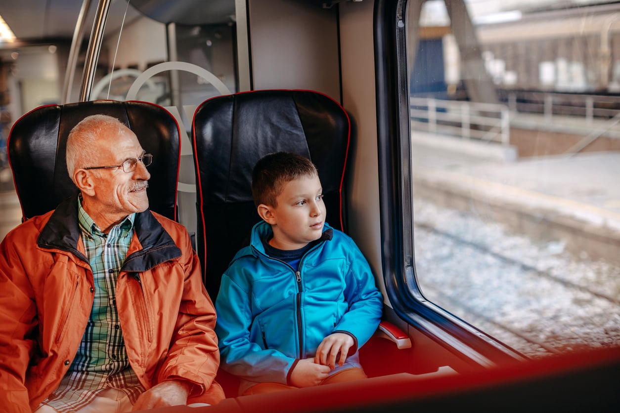 An older man and a young boy sit next to each other on a train, looking out the window. The man is smiling and wearing glasses and an orange jacket; the boy wears a blue jacket and looks thoughtful.