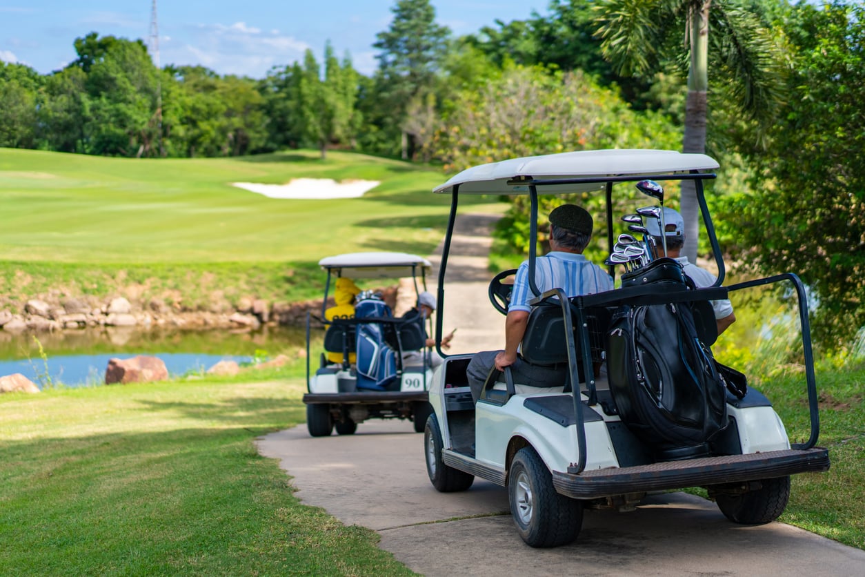 Two people ride in a golf cart on a path, following another cart on a golf course with green grass, trees, and a sand trap visible in the background.