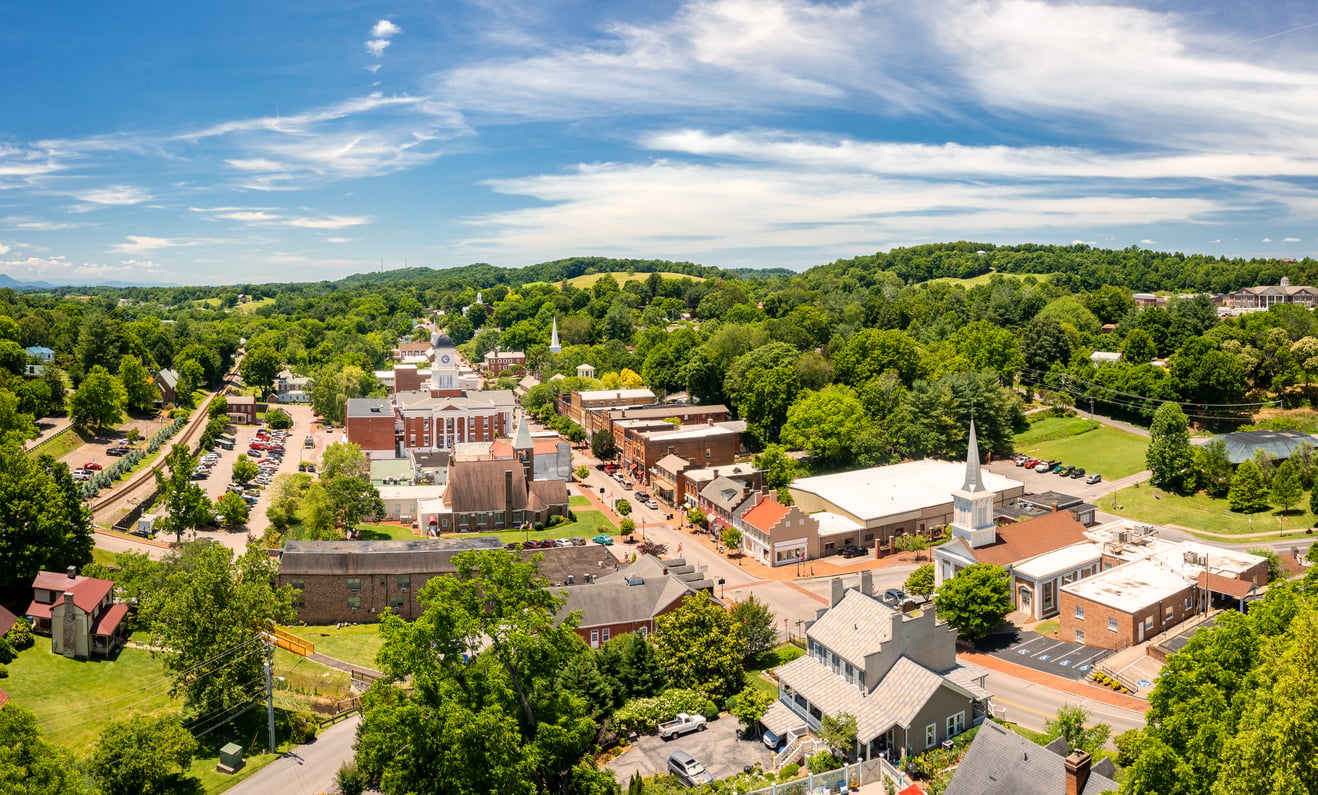 Aerial view of a small town with tree-lined streets, historic buildings, a church with a steeple, and rolling green hills under a bright blue sky with scattered clouds.