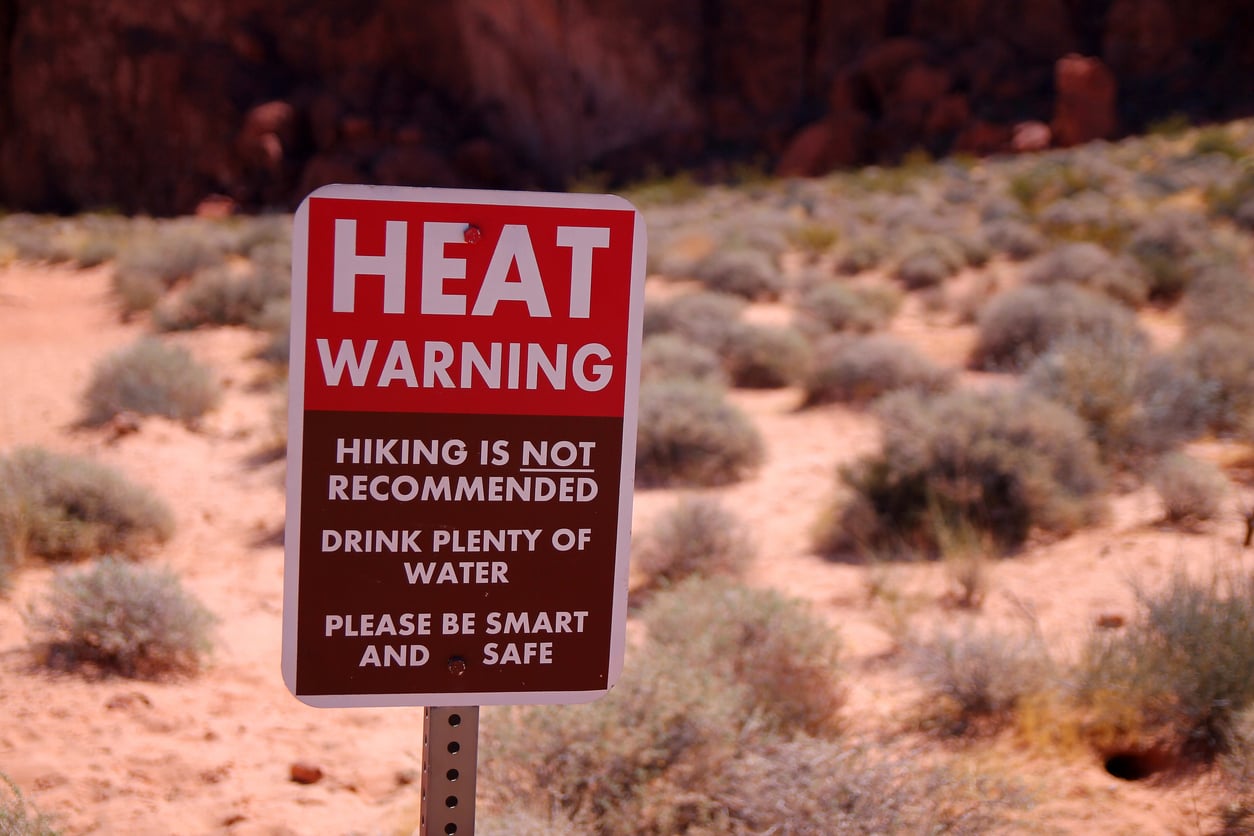 A sign in a desert area reads: “HEAT WARNING. Hiking is not recommended. Drink plenty of water. Please be smart and safe.” Dry shrubs and sand are visible in the background.