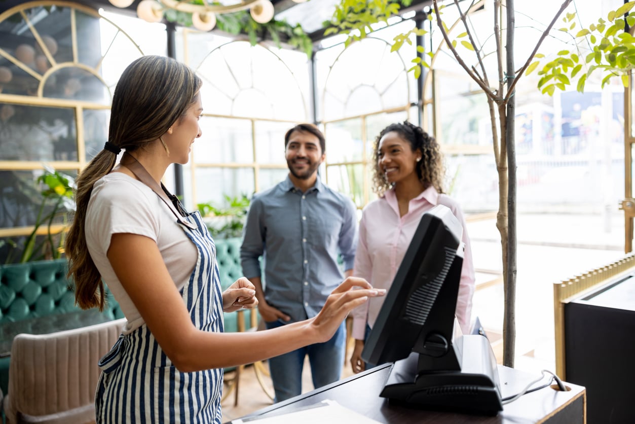 A smiling hostess at a restaurant greets a happy couple—determined to avoid common vacation mistakes—standing at the reception desk near a computer in a bright, plant-filled space.