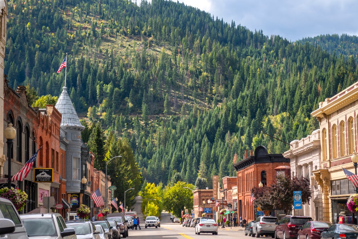 A vibrant small town street with historic brick buildings, cars parked along both sides, and green forested hills rising in the background under a partly cloudy sky.