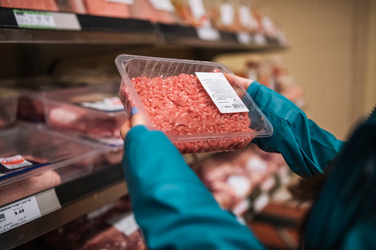 A person in a teal jacket holds a plastic-wrapped package of ground meat with a label, examining it in front of a grocery store meat section.