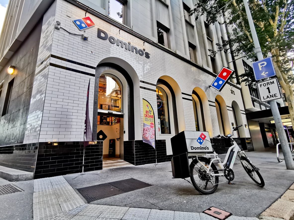 A Domino’s Pizza storefront on a city street with a branded delivery bike parked outside. The building has white and black tiles, large arched windows, and Domino’s signs visible.